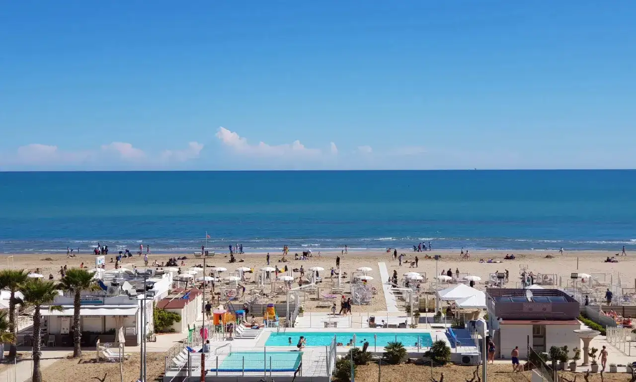 La spiaggia del cuore a Riccione, con il mare azzurro, ombrelloni, lettini, una piscina e gente che si gode il sole.