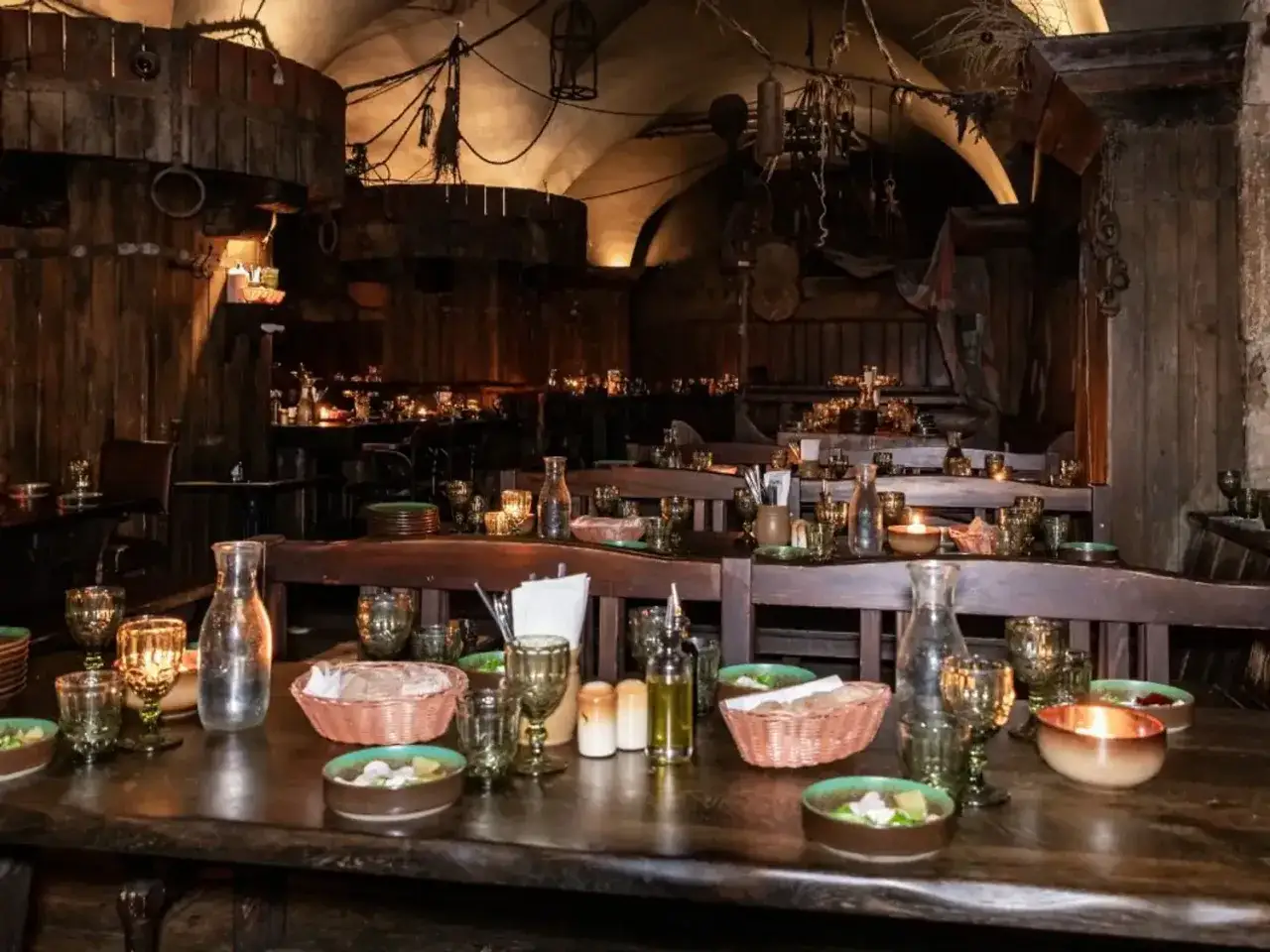 Ambiance de taverne médiévale, tables dressées avec vaisselle rustique, bougies vacillantes. Le tavernier moyen âge prépare le service.