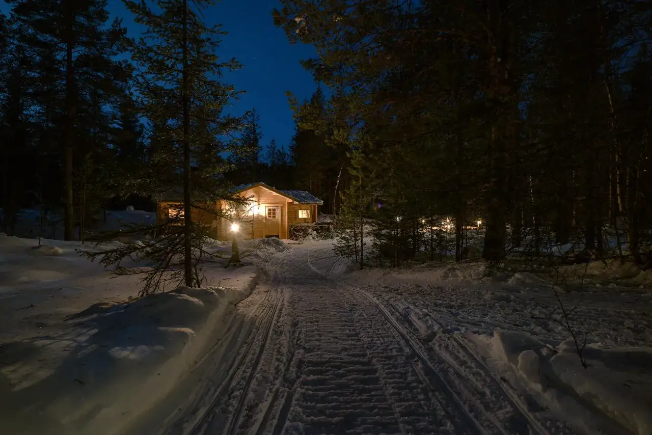 Vinterkväll vid ett mysigt vandrarhem i Jokkmokk. En snötyngd stig leder fram till en upplyst timmerstuga bland granar.