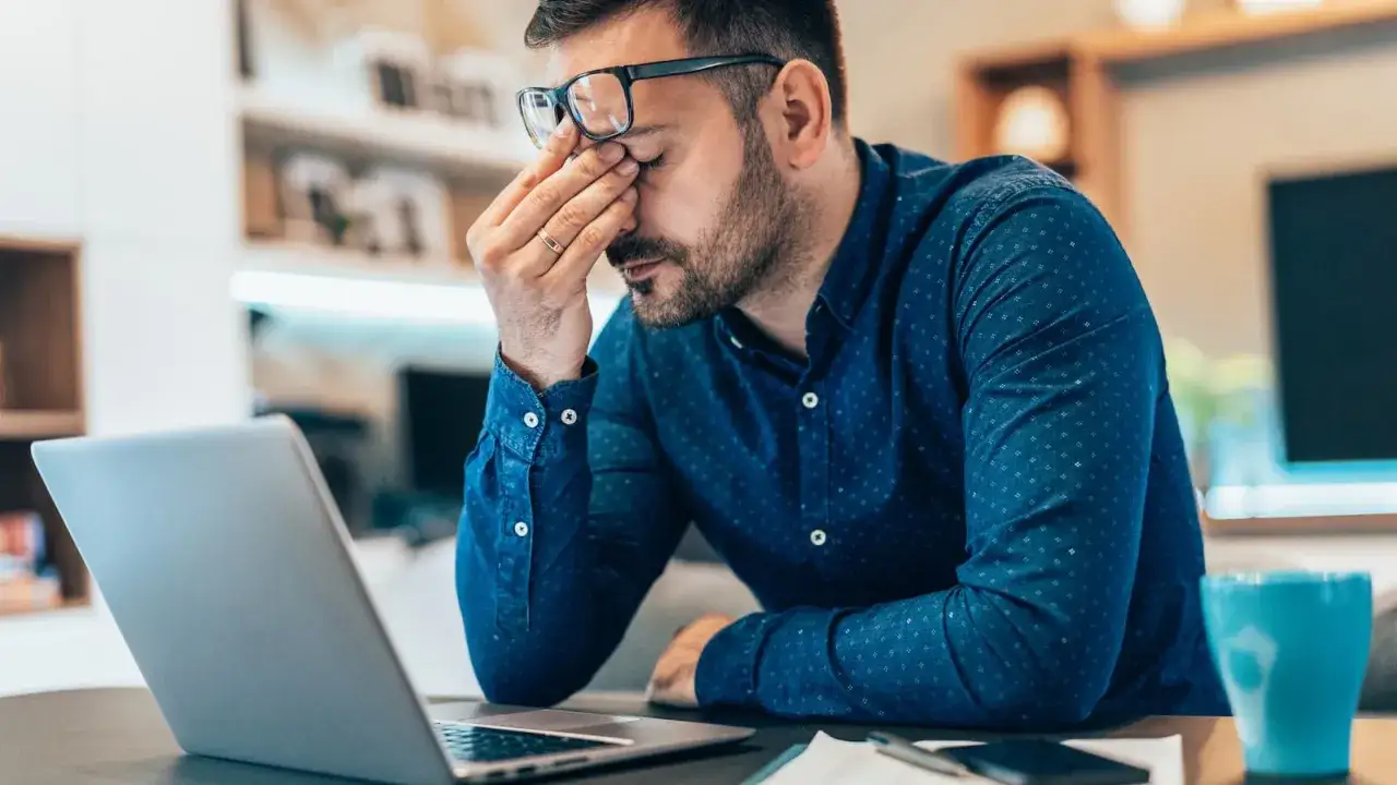 Mann reibt sich die Augen vor Frustration, fragt sich, warum ist mein Internet so langsam. Laptop und Tasse Kaffee auf dem Tisch.