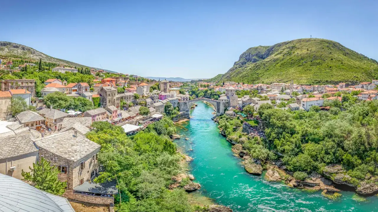 A stunning panorama of Mostar, Bosnia and Herzegovina, showcasing the iconic Stari Most bridge arching over the turquoise Neretva River. This view captures the essence of Bosnia travel, with historic stone buildings and lush green hills.