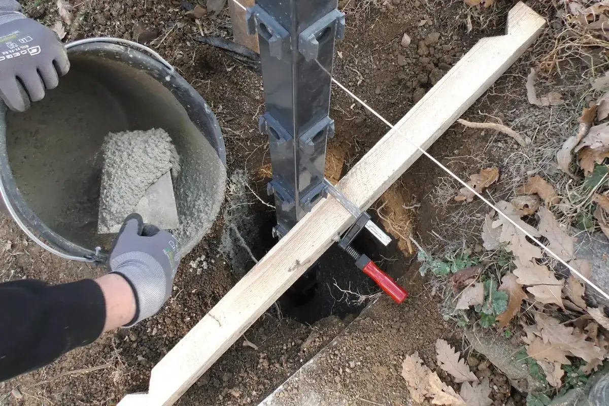 Beim Zaunpfosten einbetonieren wird Beton mit einer Kelle aus einem Eimer gesch&ouml;pft und in das Erdloch gef&uuml;llt. Ein Holzbrett st&uuml;tzt den Pfosten.