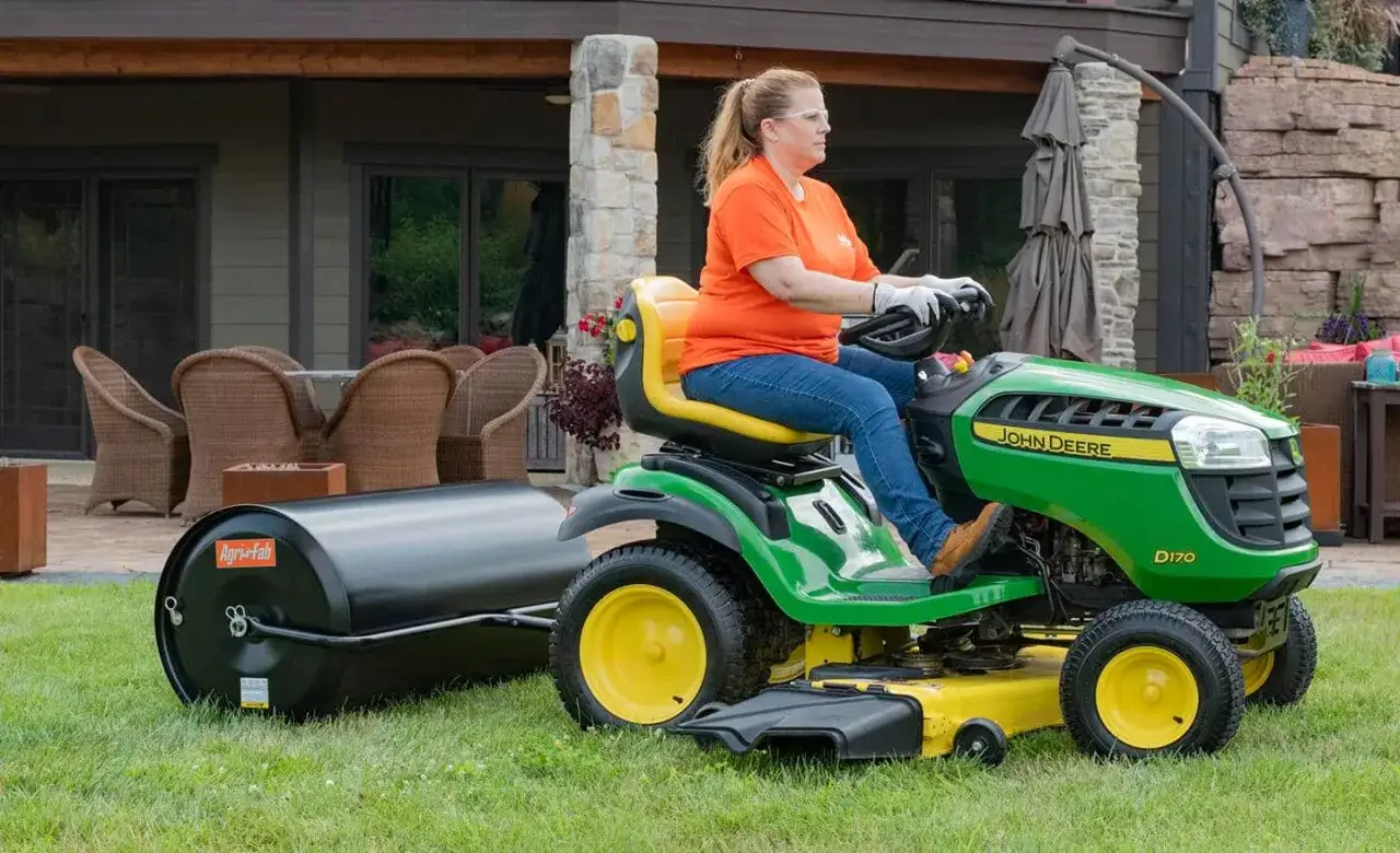 Kobieta na zielonej kosiarka do jezdzenia John Deere D170 z walcem Agri-Fab za sobą, pracuje w ogrodzie.