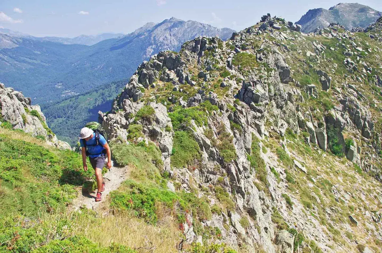 Un randonneur avec un sac à dos progresse sur un sentier rocailleux, dominant une vallée verdoyante. Le paysage montagneux est spectaculaire.