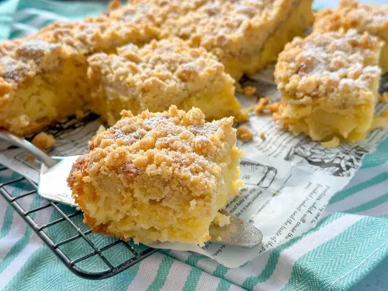 Saftiger Apfelkuchen vom Blech mit knuspriger Streuseldecke, bestäubt mit Puderzucker. Ein Stück wird gerade serviert.