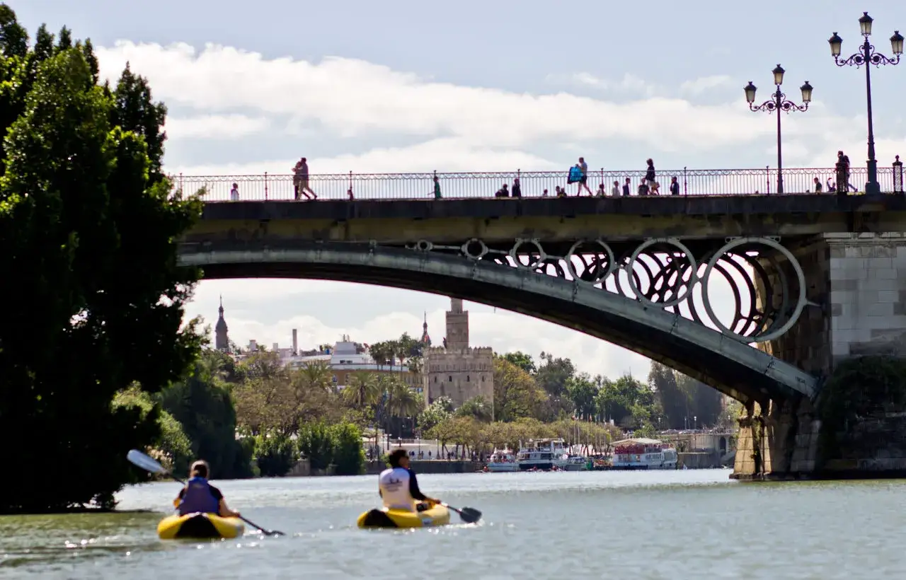 Zwei Kajakfahrer gleiten auf dem Guadalquivir, dem sevilla fluss, unter einer Br&uuml;cke mit Menschen und der Skyline von Sevilla im Hintergrund.