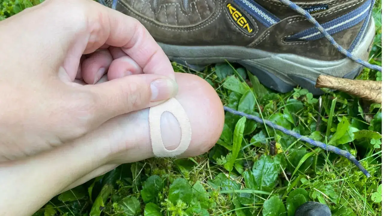 A hand applies a bandage to a toe, treating blisters from hiking boots. The shoe rests on wet grass.