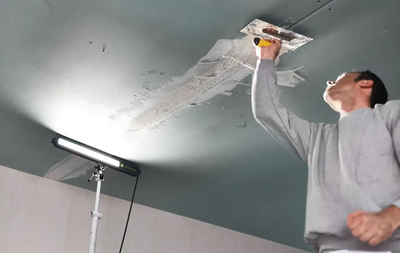 Un artisan applique de l'enduit au plafond, éclairé par une lampe rasante Festool qui révèle chaque imperfection.
