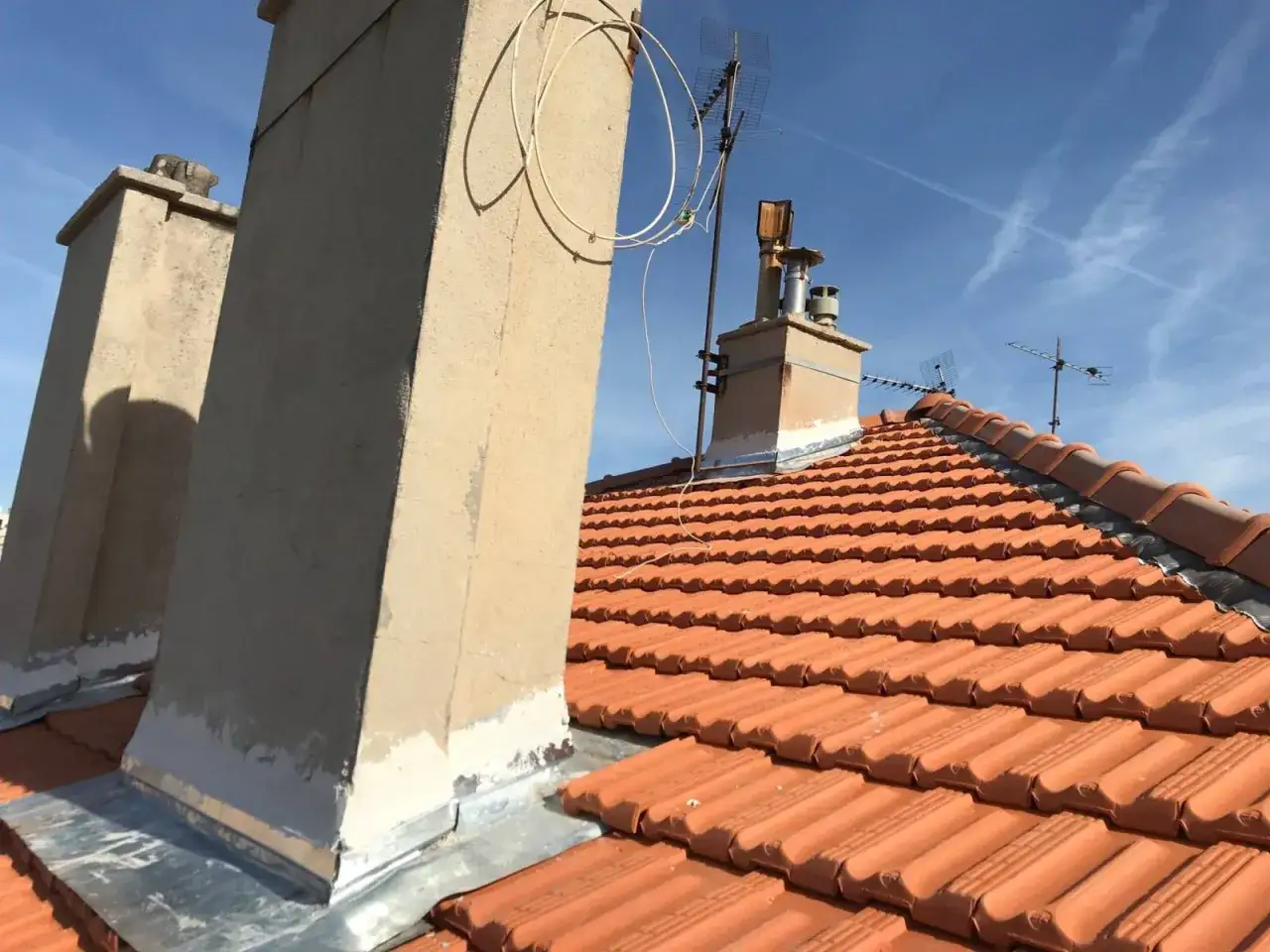 Travaux de r&eacute;novation sur un toit en tuiles rouges, avec une chemin&eacute;e en cours de d&eacute;molition et des antennes TV.