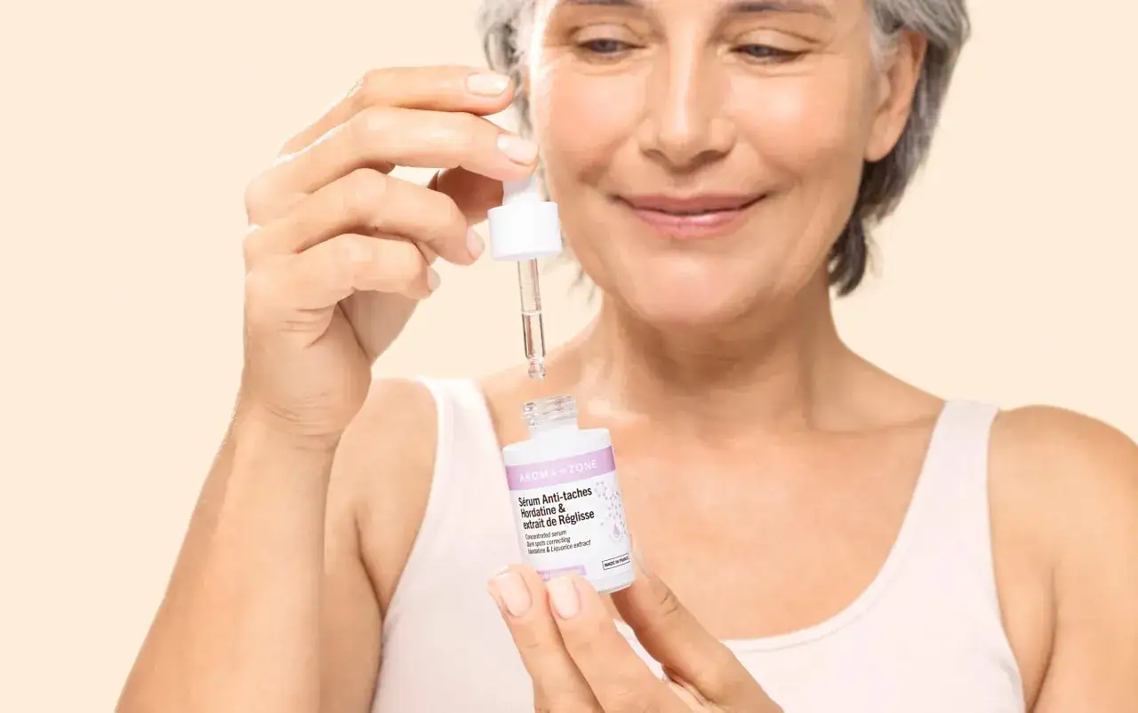 Une femme souriante tient un flacon de s&eacute;rum anti-taches &agrave; l'extrait de r&eacute;glisse, pr&ecirc;t &agrave; l'appliquer.