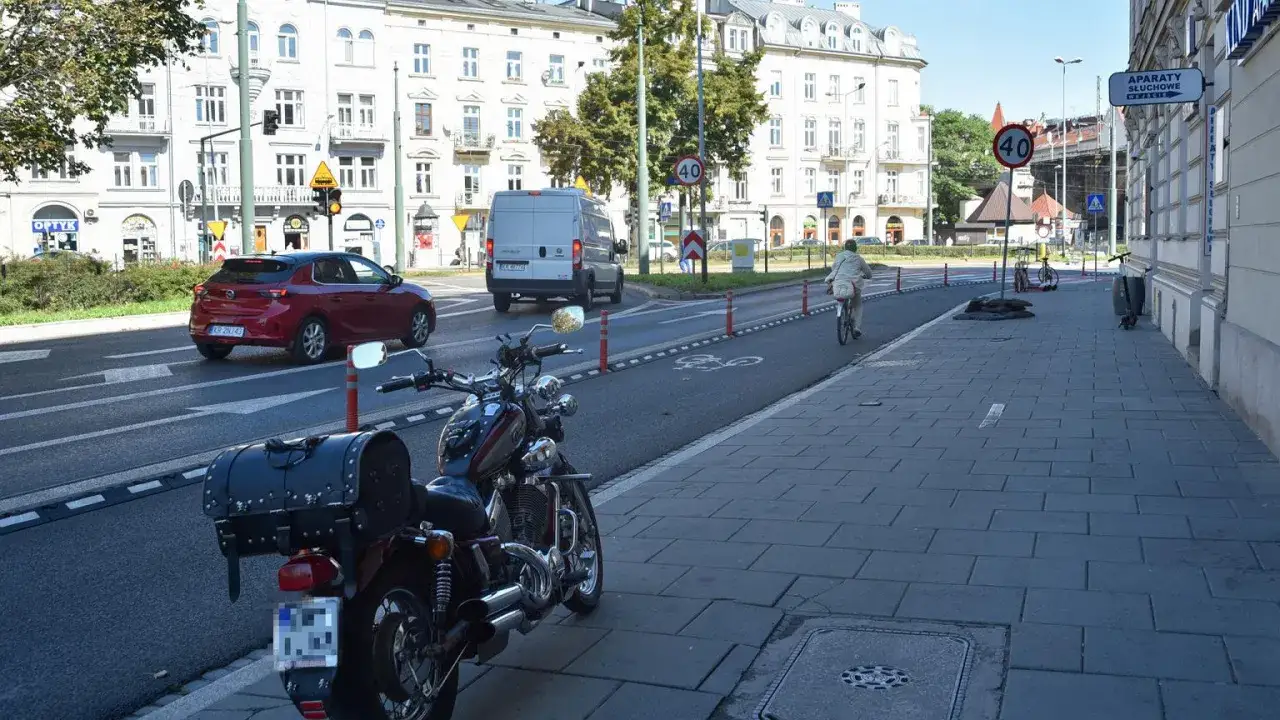 Motocykl czeka na swojego właściciela na **parkingu dla motocykli** obok ścieżki rowerowej. W tle ruch miejski.
