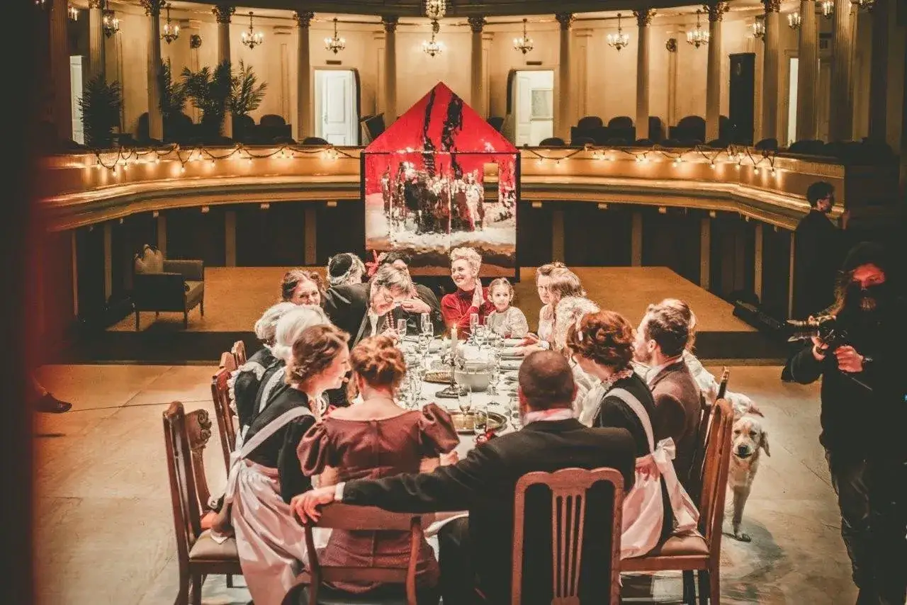 Scena z bankietem w teatrze im. H. Modrzejewskiej. Ludzie w strojach z epoki siedzą przy stole, w tle czerwona scenografia.