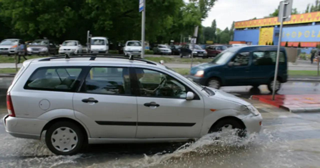Srebrny kombi jedzie przez głęboką kałużę, powodując **aquaplaning**. Woda rozpryskuje się spod kół.