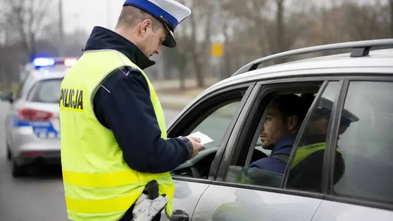 Policjant w kamizelce odblaskowej wręcza kierowcy dokument. W tle radiowóz z włączonymi sygnałami. Kierowca może się spodziewać, że zabrane prawo jazdy za prędkość to realna konsekwencja.