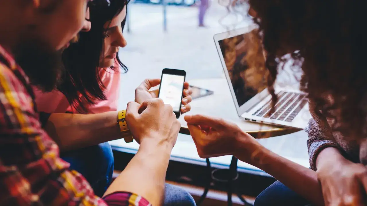 Tre v&auml;nner delar en mobiltelefon, visar positiva saker med sociala medier. En laptop st&aring;r bredvid.