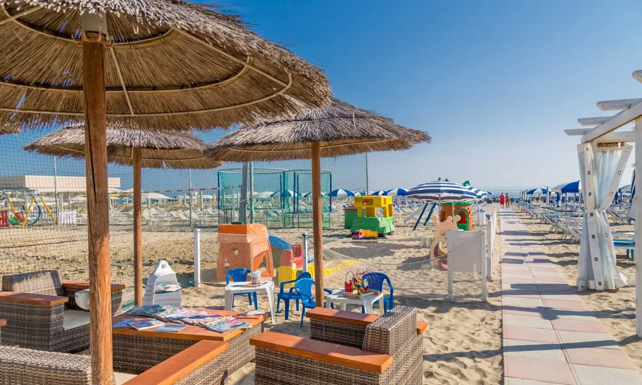 Ombrelloni di paglia e lettini sulla spiaggia di Porto Verde, con giochi per bambini e un percorso pedonale che porta verso il mare.