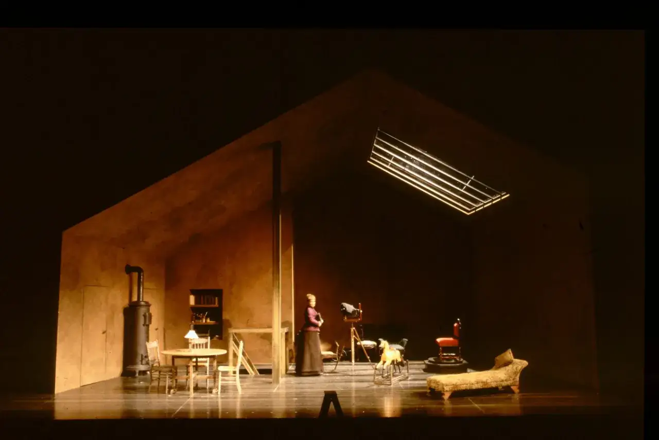 Scena teatralna z kobietą, piecem, stołem i krzesłami. W tle widać aparat fotograficzny, a na pierwszym planie leżankę. Jakby z okna wpadł promień światła, niczym dzika kaczka przelatująca przez pokój.
