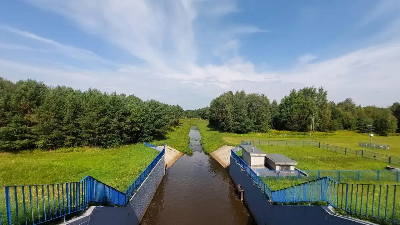 Kanał wodny prowadzący do jeziora Maziarnia, otoczony zielenią i lasem, pod błękitnym niebem z chmurami.