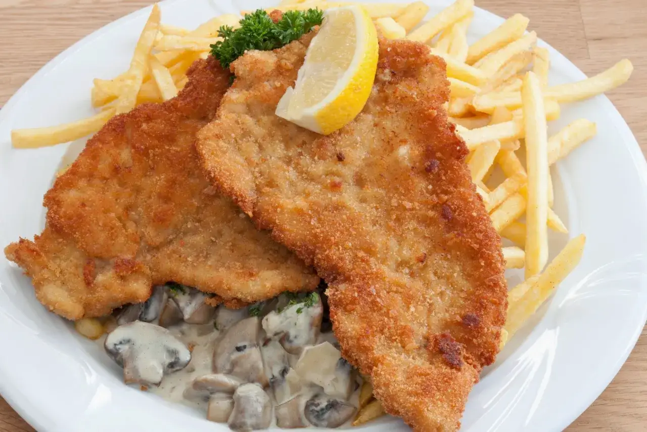 Dos milanesas de ternera doradas, acompa&ntilde;adas de papas fritas crujientes, champi&ntilde;ones en salsa cremosa y un gajo de lim&oacute;n.