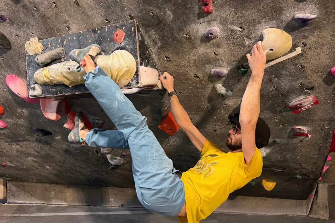 Mężczyzna w żółtej koszulce wspina się po ścianie z chwytami. To właśnie bouldering – wspinaczka bez liny na niskich ścianach.