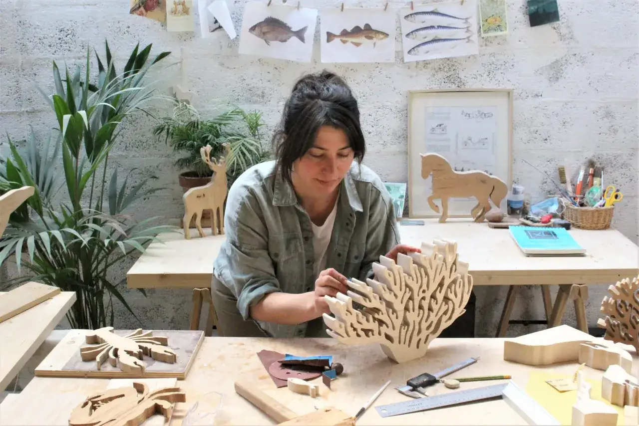 Une artiste concentr&eacute;e travaille dans son atelier, entour&eacute;e de ses cr&eacute;ations en bois. Elle semble sculpter le bois avec soin, donnant vie &agrave; des formes marines et animales.