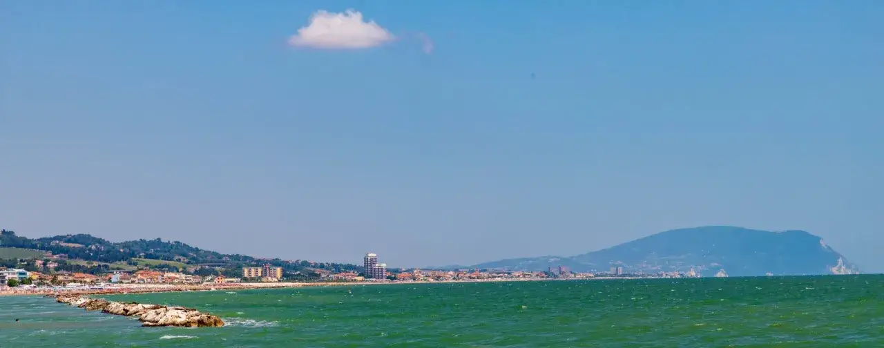 Mare verde e cielo azzurro con una nuvola solitaria sopra la costa di Porto Civitanova, con edifici e colline sullo sfondo.
