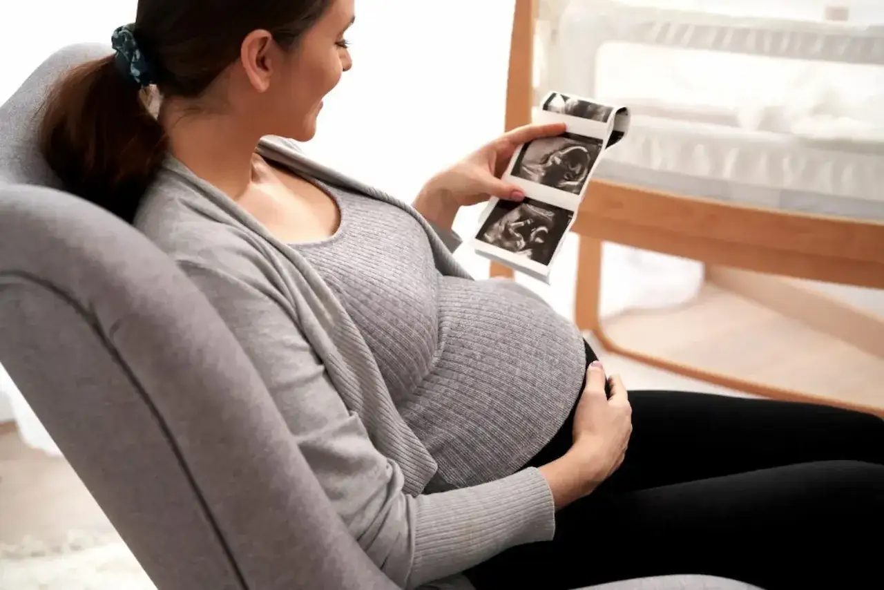 Szczęśliwa przyszła mama ogląda zdjęcia z usg ciąży, trzymając je w dłoni. W tle widać łóżeczko dziecięce.