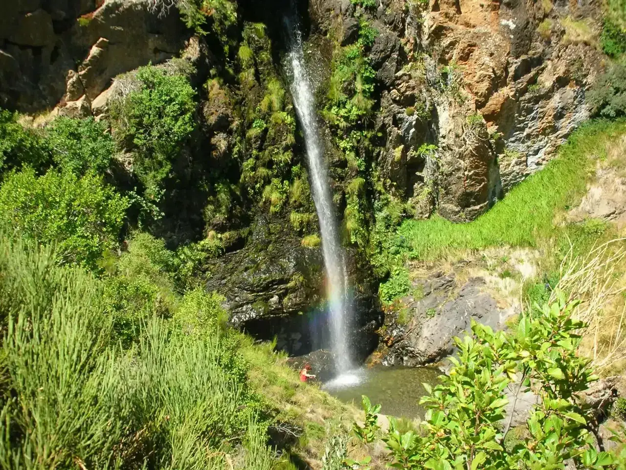 Una persona observa la cascada de Mazobre, con un arco&iacute;ris formado por el roc&iacute;o. Vegetaci&oacute;n exuberante rodea la ca&iacute;da de agua.