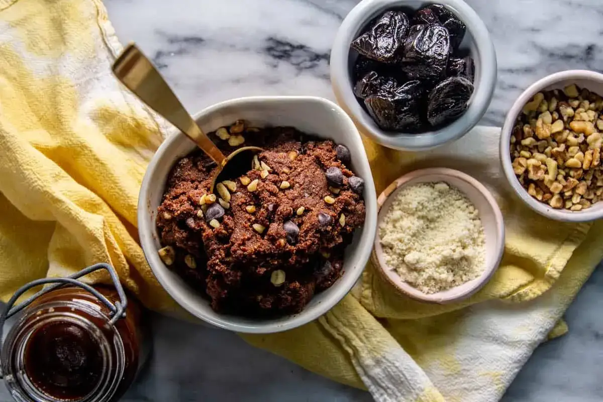 Chokladig cookie dough recept med n&ouml;tter och chokladknappar. Ingredienser som katrinplommon, valn&ouml;tter och mandelmj&ouml;l syns bredvid.