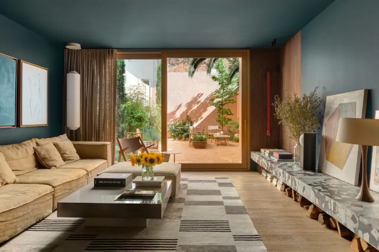 Ein langes, schmales Wohnzimmer mit beiger Couch, modernem Couchtisch und Teppich. Durch die Glastür blickt man in einen sonnigen Innenhof.