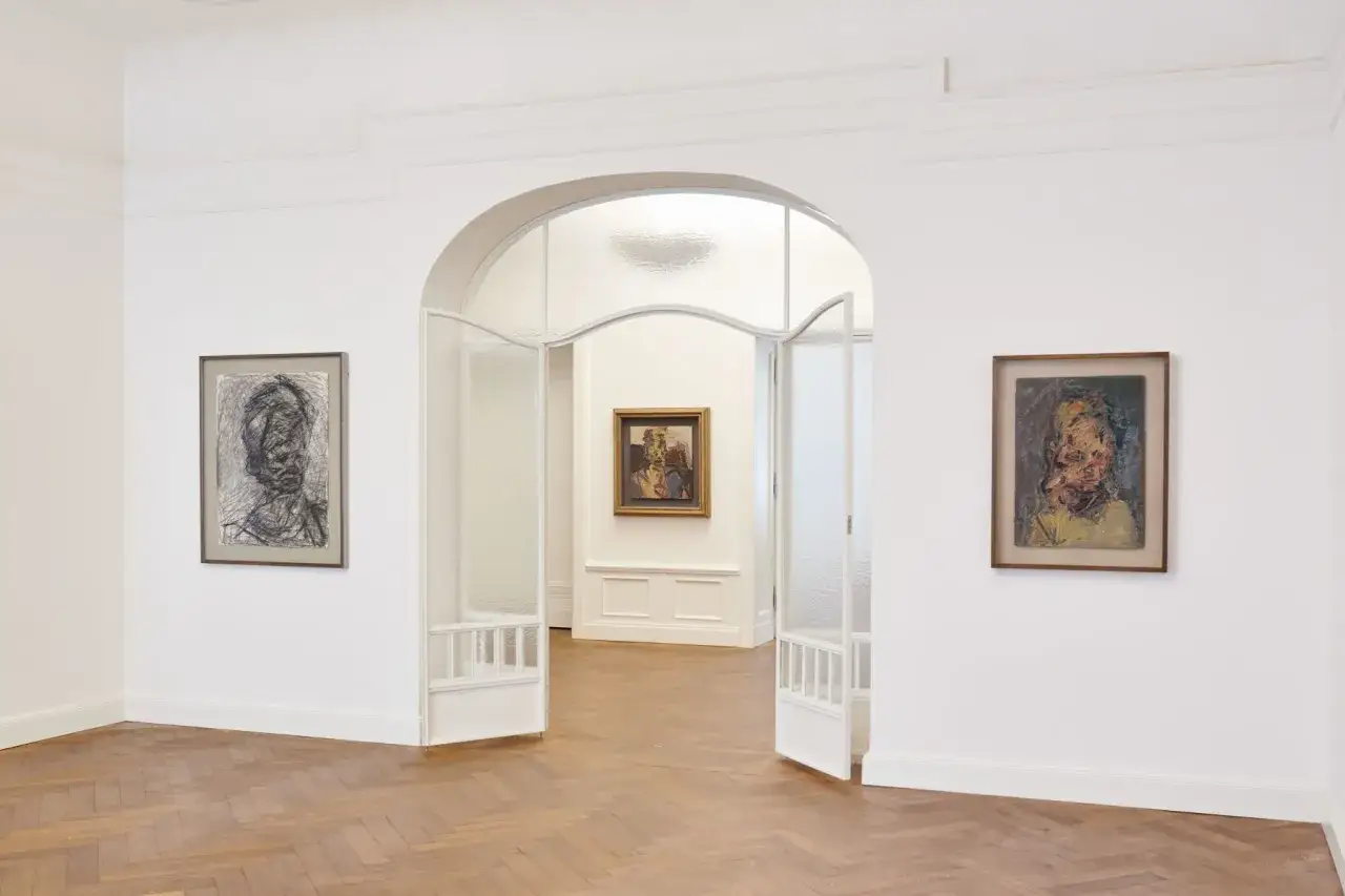 Portraits on display at Michael Werner Gallery, one a charcoal sketch, another a vibrant oil painting, draw viewers through an arched doorway.