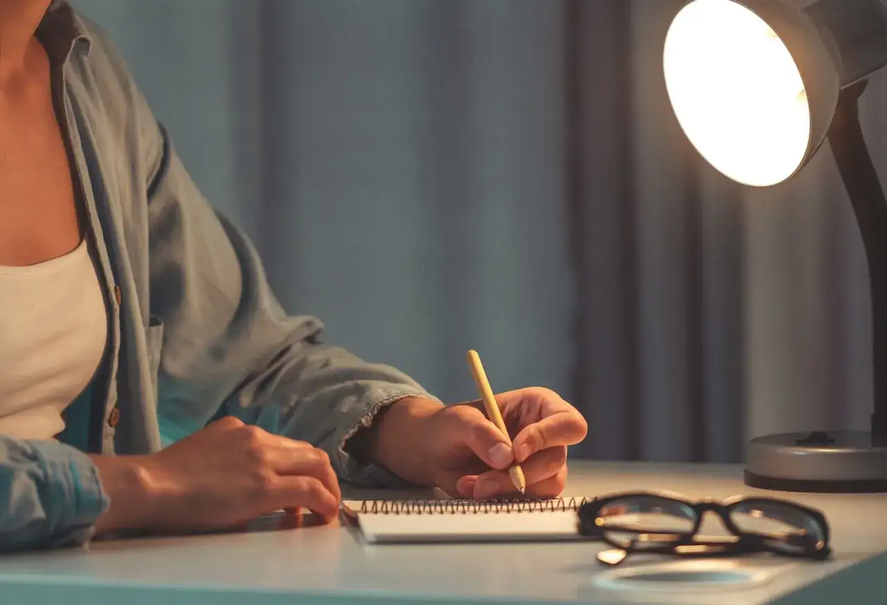 A person writes in a notebook under a desk lamp, perhaps processing common insecurities.