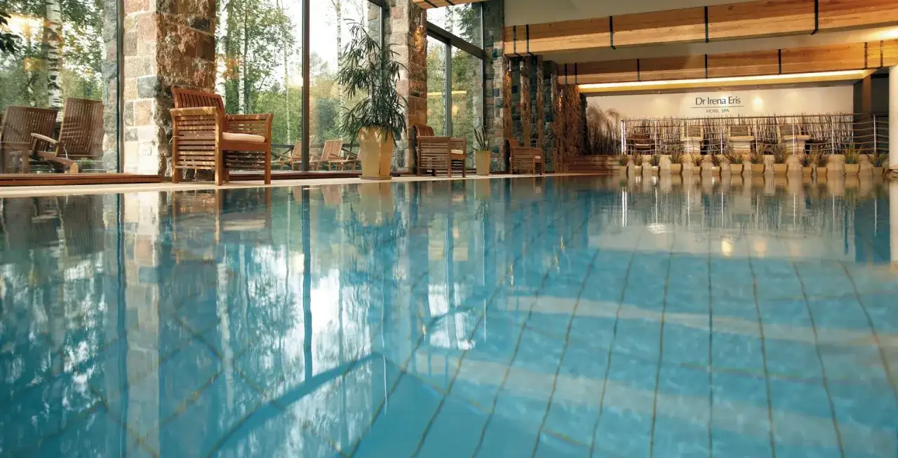 Basen w hotelu Dr Irena Eris Hotel Spa. Drewniane leżaki i rośliny w donicach przy basenie z błękitną wodą.