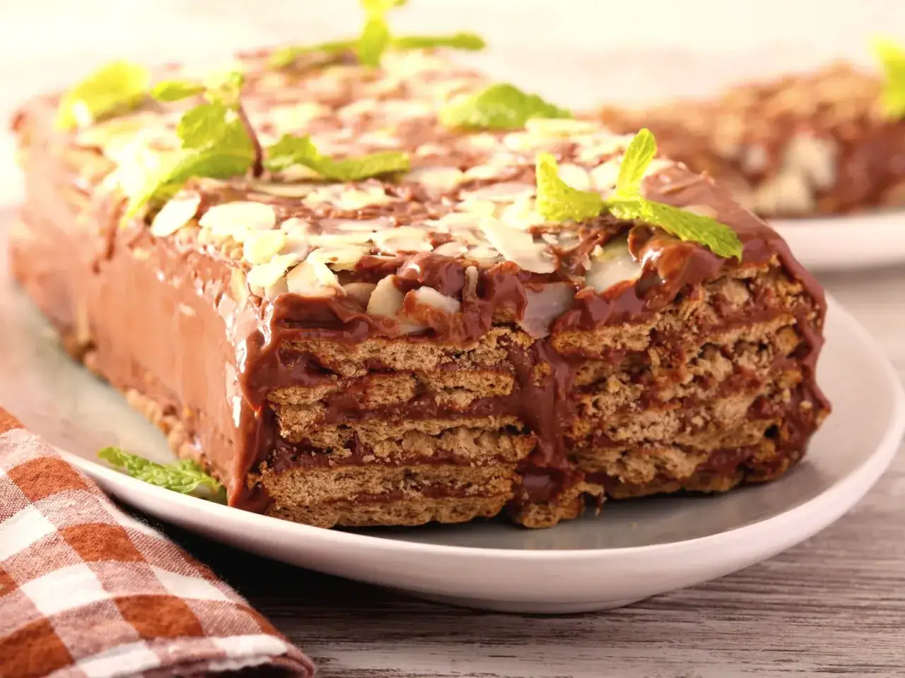 Tanie ciasto bez pieczenia z herbatnikami, oblane czekoladą i posypane płatkami migdałów, ozdobione listkami mięty.