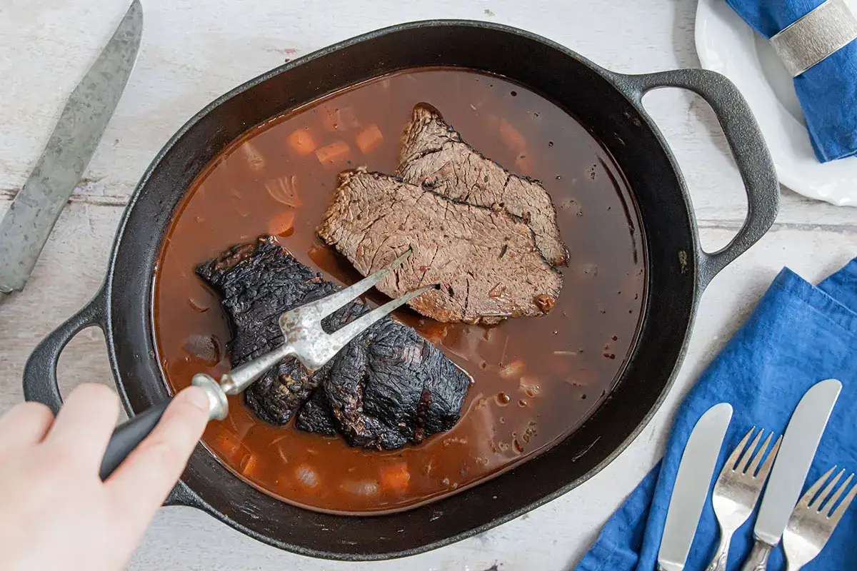 Zwei Scheiben fertig eingelegter Sauerbraten in dunkler Sauce in einer Gusseisenpfanne. Ein Fleischgabel hält ein Stück.