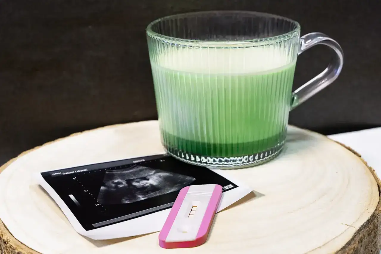 Kubek zielonego matcha latte obok pozytywnego testu ciążowego i zdjęcia USG. Czas na zdrowe napoje w ciąży.