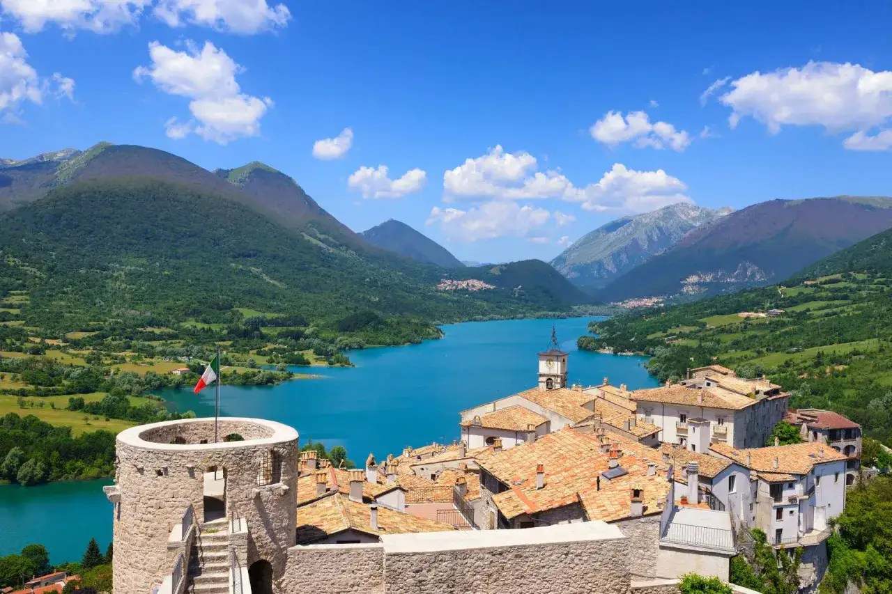 Villetta Barrea: un borgo con torre medievale e bandiera italiana domina un lago turchese, circondato da montagne verdi. Cosa vedere in questo incantevole paesaggio.