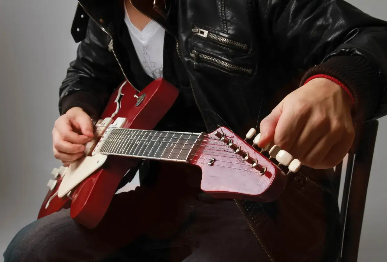 Mężczyzna w skórzanej kurtce nastroił czerwoną gitarę elektryczną, obracając klucze na główce.