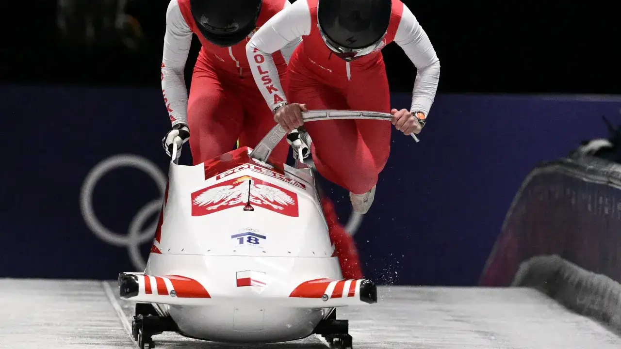 Klaudia Adamek i jej partnerka w biało-czerwonych strojach z napisem POLSKA pędzą w d&oacute;ł lodowego toru w bobslejach.