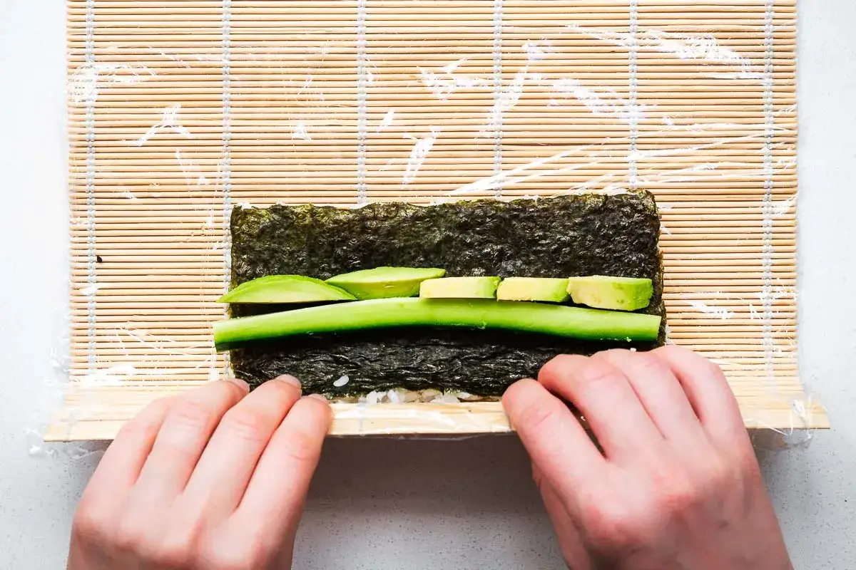 Ręce zwijają **uramaki** na macie bambusowej. Na nori ułożono ryż, ogórek i awokado.