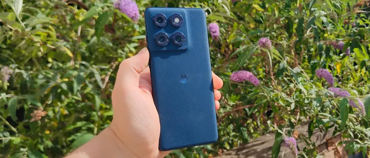 En hand h&aring;ller en m&ouml;rkbl&aring; Motorola Edge 60 Fusion med fyra kameror mot en bakgrund av gr&ouml;na blad och lila blommor.