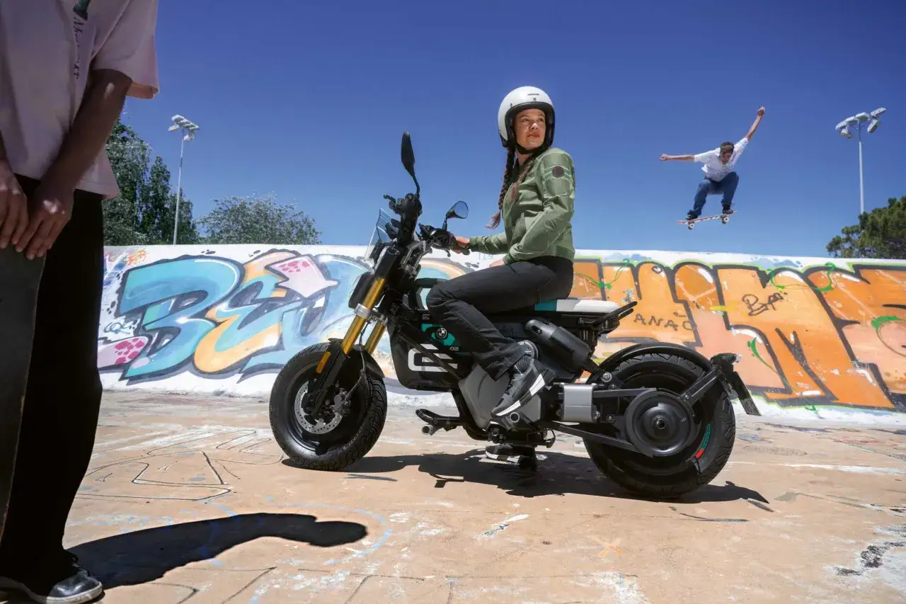 Dziewczyna na motocyklu BMW CE02 w skateparku, podczas gdy skater wykonuje skok w tle.