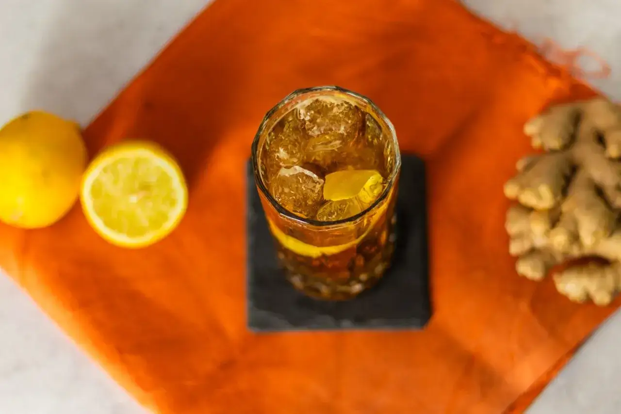 Ein Glas mit Eis und einem Zitronenschnitz, daneben Zitronen und Ingwer. Die Farbe erinnert an den Hals eines Pferdes.