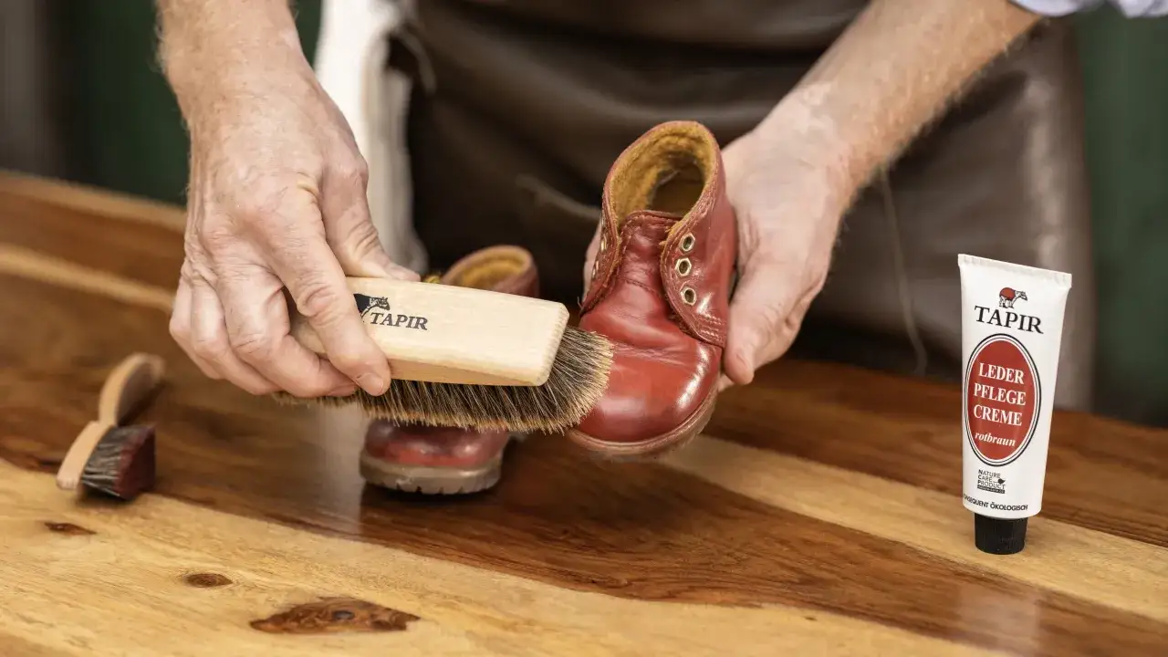 Mains expertes appliquant une cr&egrave;me pour nourrir chaussure cuir naturellement. Brosse et tube de cr&egrave;me Tapir sur un &eacute;tabli en bois.