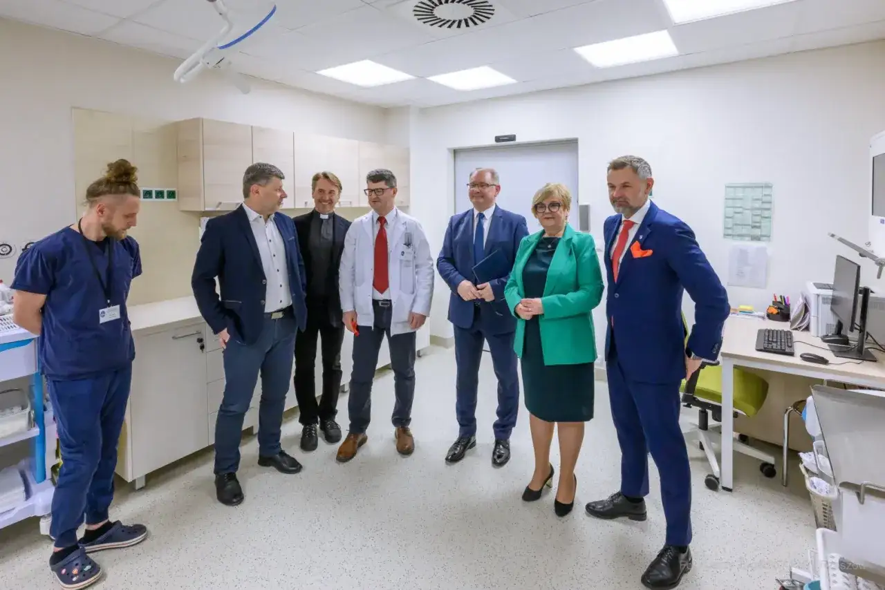 Grupa osób w strojach medycznych i garniturach stoi w nowoczesnym gabinecie lekarskim.