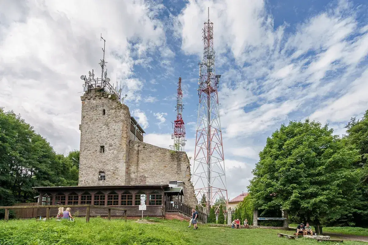 Kamienna wieża i maszty telekomunikacyjne na szczycie Chełmiec. Ludzie odpoczywają na trawie.