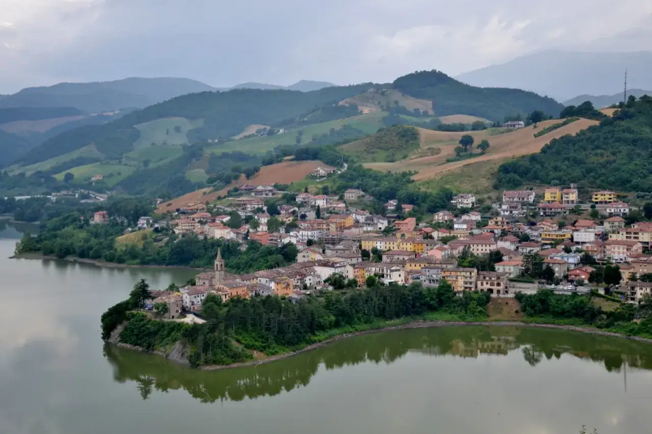 Borgo pittoresco affacciato su un lago, con case colorate e colline verdi sullo sfondo, tipico della regione di Urbino.