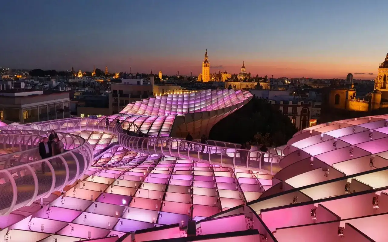 The Metropol Parasol, known as Las Setas de Sevilla, glows with pink and purple lights at dusk, overlooking the historic cityscape.