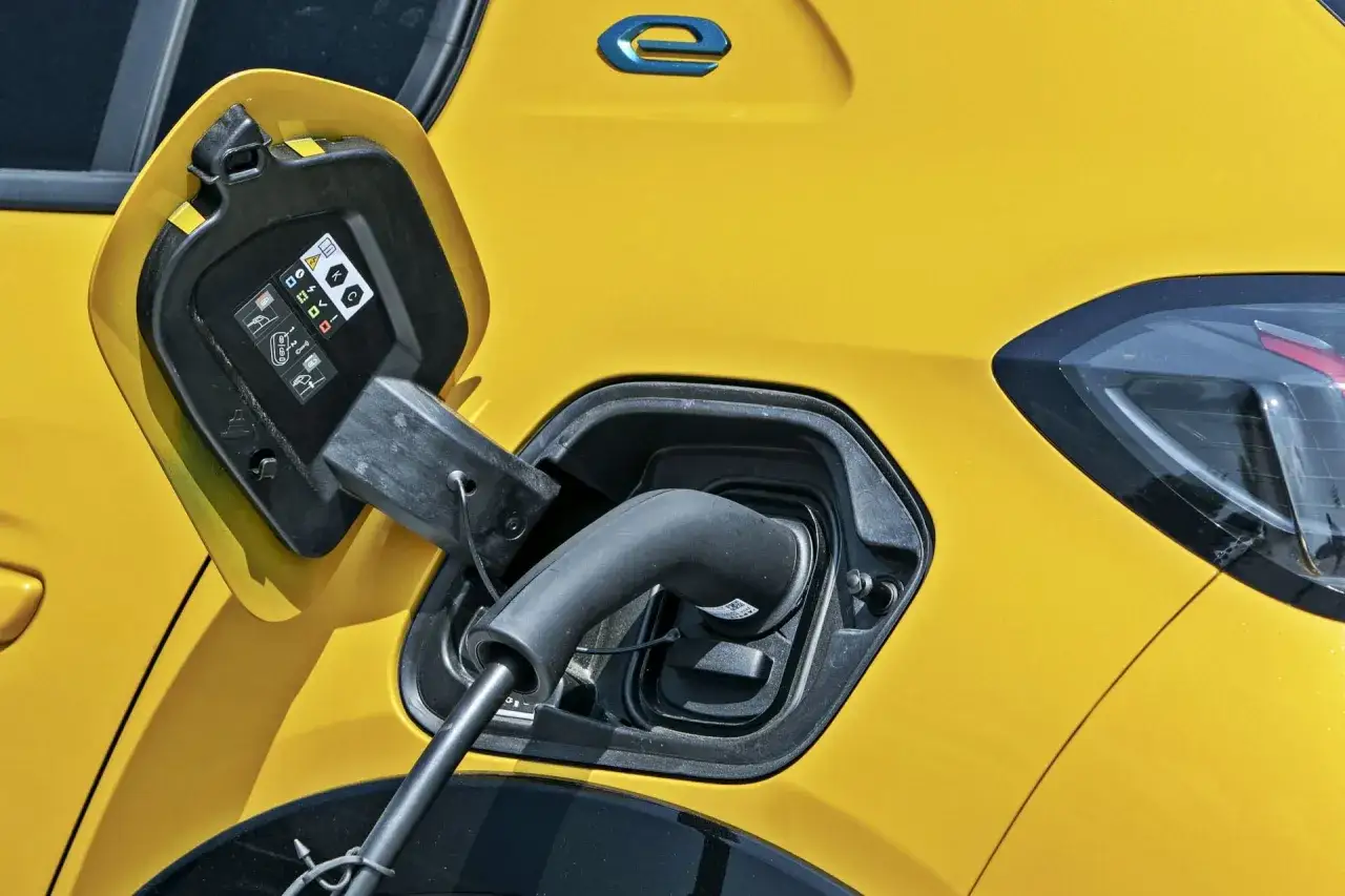Żółty Peugeot e-208 jest ładowany. Widać otwartą klapkę gniazda ładowania z kablem.