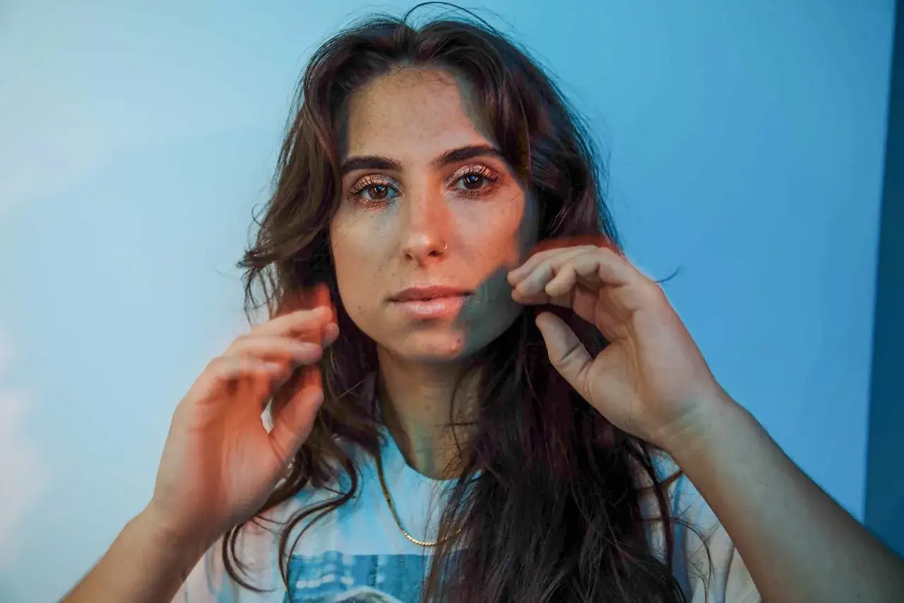 Alysa Vanderheym, with her expressive eyes and freckled skin, is framed by warm light and a cool blue background. Her hands gently touch her face, creating a sense of introspection.